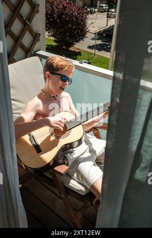 Un ragazzo siede comodamente su una sedia a sdraio su un balcone, suona una chitarra classica e indossa gli occhiali da sole in una giornata di sole. Foto Stock