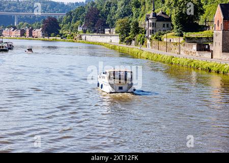 La Mosa con vista sul retro della barca a vela sulle acque calme, alla periferia della città di Dinant, agli edifici e alla strada di campagna lungo il fiume sullo sfondo, in estate soleggiata Foto Stock