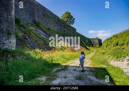 Turista femminile in piedi di profilo su un sentiero che guarda in alto, tra imponenti alte mura di pietra alla fortezza di Charlemont, cielo blu sullo sfondo, soleggiato summ Foto Stock