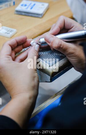 Dall'alto un primo piano irriconoscibile di mani che intagliano meticolosamente un piccolo oggetto con uno strumento rotante nello spazio di lavoro di un gioielliere, concentrato e detai Foto Stock