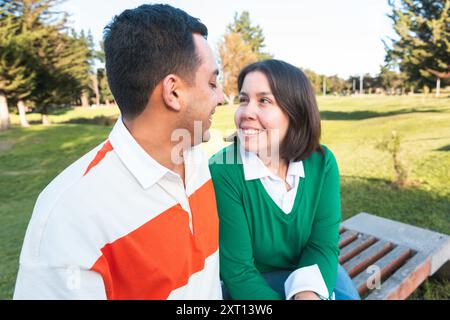 Una giovane e gioiosa coppia latina siede da vicino su una panchina del parco, condividendo un sorriso in un ambiente naturale e rilassato Foto Stock