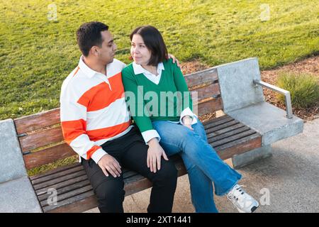 Una giovane coppia latina siede da vicino su una panchina del parco, condividendo un momento di affetto con sorrisi e uno sguardo tenero in un ambiente tranquillo all'aperto Foto Stock