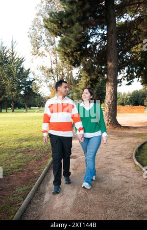 Una giovane coppia latina si gode una piacevole passeggiata attraverso un parco verdeggiante, condividendo un momento di connessione mentre camminano mano nella mano, con alberi alti nel Foto Stock