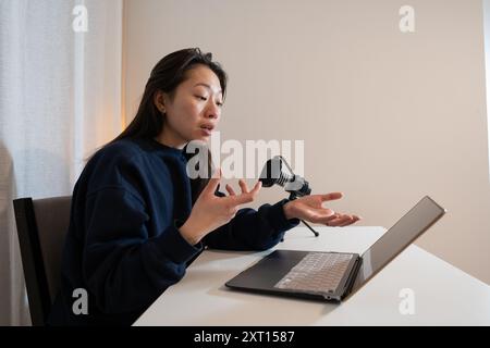 Una donna concentrata esegue gesti mentre registra contenuti audio con un microfono e un computer portatile in un ambiente domestico Foto Stock