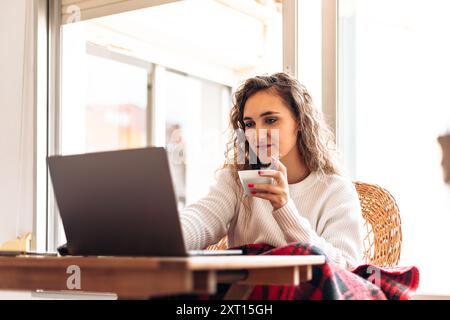 Giovane freelance che si gode il tè e lavora su un netbook mentre siede al tavolo a casa durante un progetto remoto in un appartamento moderno Foto Stock