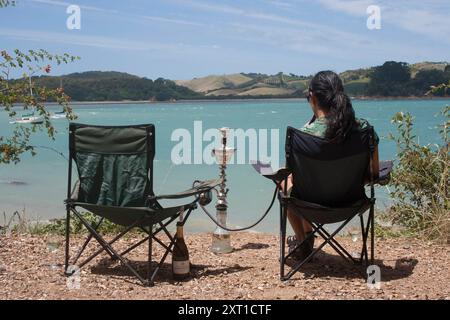 Una donna si rilassa davanti ad una tranquilla vista del lago, seduto su una sedia portatile con un allestimento a narghilè e una bottiglia di vino Waiheke Island New Zealand evpe00452 Copyright Foto Stock