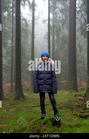 Una persona si trova su un terreno coperto di muschio in una foresta nebbiosa e indossa un cappotto blu navy con cappuccio foderato in pelliccia e un berretto blu brillante. Bola02440 Copyrigh Foto Stock