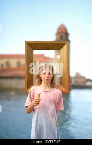 Un bambino sorridente che tiene in mano un cono gelato si erge dietro una cornice vuota con un edificio storico e acqua sullo sfondo. Bola02457 Copyright Foto Stock