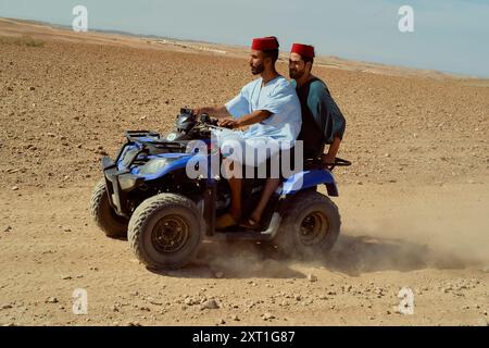 Due uomini in abbigliamento tradizionale che si godono un giro in quad attraverso un paesaggio desertico. Bola02518 Copyright: XConnectxImagesx Foto Stock