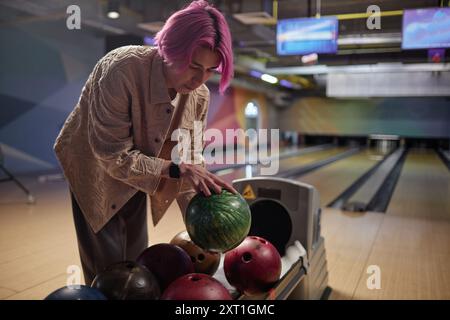 Persona con i capelli rosa nel bowling che seleziona la palla da bowling da rack circondata da un ambiente vivace e colorato con corsie sullo sfondo Foto Stock