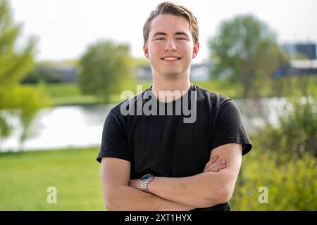 Portret van een giovane millenario sorridente in piedi all'aperto con le braccia incrociate di fronte a un corpo d'acqua naturale sfocato e al verde Molenhoek Nederl Foto Stock