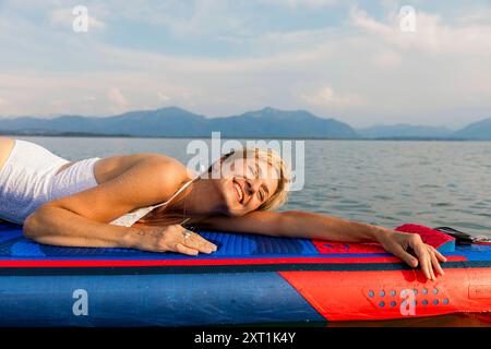 Donna sorridente sdraiata su un paddleboard che galleggia su acque calme con montagne in lontananza. Stef00067 Copyright: XConnectxImagesx Foto Stock