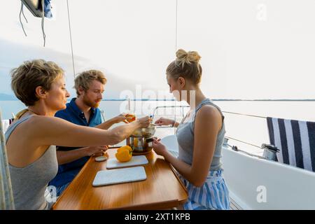 Tre persone si godono un pasto insieme sul ponte di una barca a vela con un tranquillo sfondo d'acqua durante il tramonto. Stef00110 Copyright: XConnectxImagesx Foto Stock
