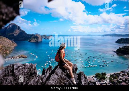 Una donna siede in cima a una cima rocciosa che si affaccia su una vista costiera mozzafiato con barche e isole sotto un cielo azzurro. Filippine rist00026 Copyright: Foto Stock