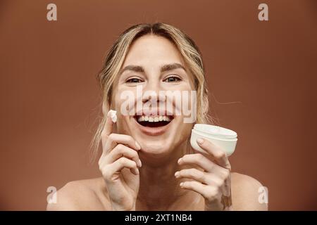 Una giovane donna con vitiligine sorride brillantemente mentre applica l'idratante sul viso, mostrando la sua pelle bella e unica. Foto Stock