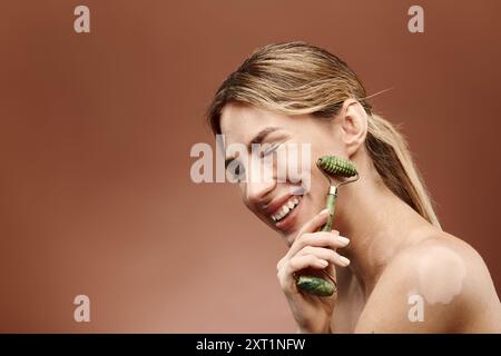 Una giovane donna con vitiligine sorride mentre usa un rullo di giada per massaggiarsi il viso, mostrando la sua routine per la cura della pelle. Foto Stock