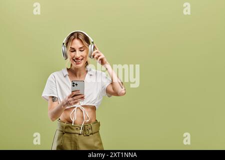 Una donna con vitiligine sorride con le cuffie su sfondo verde, indossa camicia bianca e pantaloni kaki, usando il suo telefono. Foto Stock