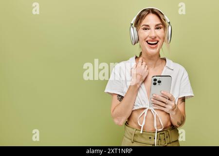 Una giovane donna che indossa un top bianco, pantaloni kaki e cuffie sorride brillantemente mentre tiene in mano uno smartphone. Foto Stock