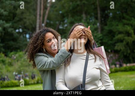 La donna sorprende la sua amica coprendo scherzosamente gli occhi in un parco lussureggiante, entrambi sorridenti. Panc05437 Copyright: XConnectxImagesx Foto Stock