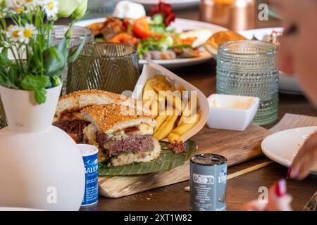 Primo piano di un delizioso hamburger tagliato a metà con patatine fritte e condimenti assortiti su un tavolo di legno, con una persona fuori fuoco in primo piano. panc05 Foto Stock