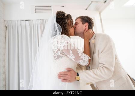 La sposa e lo sposo condividono un bacio appassionato il giorno del loro matrimonio, con lo sposo in abito beige e la sposa con un abito e un velo in pizzo bianco. panc059 Foto Stock