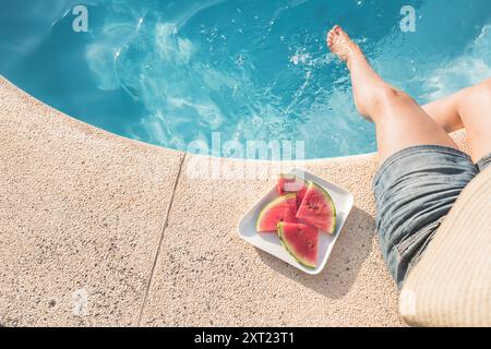 Una donna siede in piscina con le gambe in acqua, un cappello di paglia e un piatto di fette di anguria accanto a lei, godendosi una giornata estiva Foto Stock