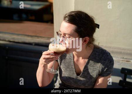 Donna in camicia a righe sorseggiando un drink da un bicchiere in un ambiente esterno con luce solare bola02807 Copyright: XConnectxImagesx Foto Stock