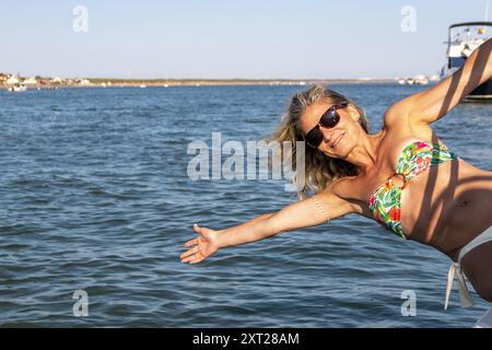Donna bionda con capelli sciolti e occhiali da sole si appoggia sul lato di una barca sopra le onde e il mare blu Foto Stock