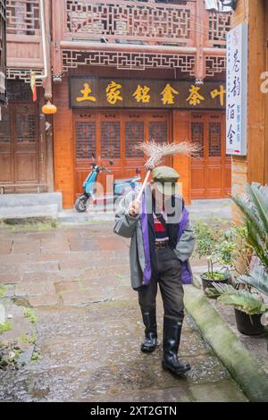 Un uomo anziano che spazia su strade di ciottoli bagnate in un tradizionale villaggio cinese, circondato da un'architettura in legno e pietra. Foto Stock