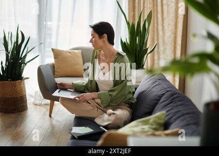 Una donna attraente lavora a distanza da casa, comodamente seduto su un divano in una stanza ben illuminata. Foto Stock