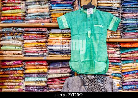 Una camicia verde è appesa davanti a scaffali impilati pieni di camicie indiane di diversi colori. Foto Stock
