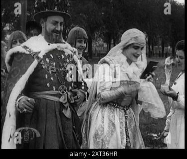 L'attrice Henny Porten vestita in costume d'epoca indossando il trucco da un compatto di polvere, mentre Emil Jannings e altri attori maschi tedeschi stanno accanto a lei, 1920 anni Riprese '...all'esterno del lotto di, di Believe IT or Not, la produzione cinematografica [muta] tedesca di Ann Boleyn. Non solo il regista Ernst Lubitsch stava facendo buoni film, ma utilizzava anche un tema britannico per la sua sceneggiatura. La star del film è l'attore Emil Jannings nel ruolo di Enrico VIII. Per Elstree, occhio per occhio, un film per un film". Da "Time to Remember - Came the Dawn", 1925 (Reel 2); sbircia all'industria cinematografica britannica di t Foto Stock