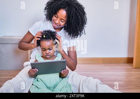 Una giovane madre afro-americana acconcia delicatamente i capelli della sua bambina mentre entrambi si impegnano con un tablet digitale in un ambiente accogliente Foto Stock