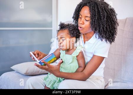 Un momento tenero come una madre nera guida la sua bambina attraverso un libro, entrambi seduti comodamente su un divano di colore chiaro Foto Stock