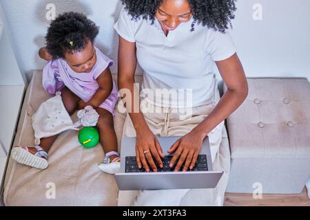 La madre afroamericana gioca in multitasking su un laptop mentre la figlia gioca con i giocattoli accanto a lei, raffigurando un ambiente multitasking che alimenta Foto Stock