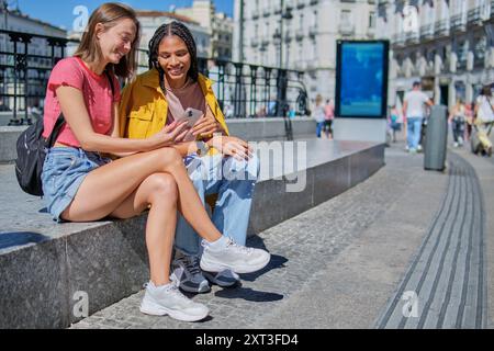 Due giovani donne multietniche condividono un momento di gioia guardando lo schermo mentre sono sedute su una panchina quadrata della città, bagnate dalla luce del sole. Foto Stock