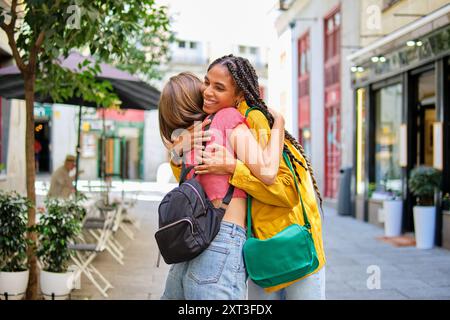Due giovani donne multietniche, una con trecce, che si abbracciano gioiosamente in una strada urbana trafficata, esprimendo amicizia e felicità mentre sorridono e si tengono in braccio Foto Stock
