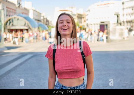 Una giovane donna, ridendo con gioia, si trova in una vivace piazza della città a guardare la macchina fotografica. È vestita con un casual top rosso con un cinturino nero sul retro Foto Stock
