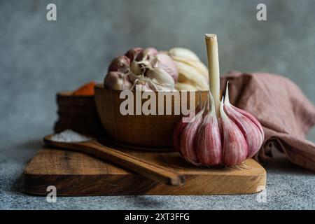 Bulbi d'aglio freschi e singoli spicchi esposti su un tagliere di legno, accompagnati da una ciotola di legno, che crea un tema di cucina visivamente ricco Foto Stock