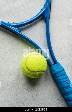 Un'immagine dettagliata di primo piano che mostra una racchetta da tennis blu con impugnatura avvolta e una palla da tennis gialla luminosa appoggiata su una superficie di cemento testurizzato Foto Stock