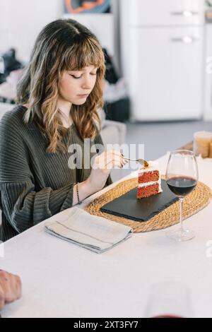Una donna che guarda in basso una fetta di torta di velluto rosso che sta per mangiare, con un bicchiere di vino rosso accanto a lei. E' seduta ad un tavolo da pranzo bianco, CA Foto Stock