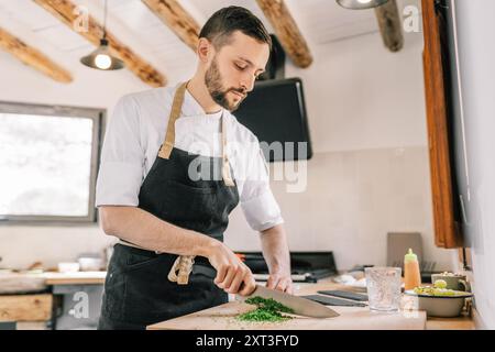 Un uomo, con una camicia bianca e un grembiule nero, si trova in una cucina casalinga, concentrata sul tritare le erbe. Lo chef sta guardando in basso con attenzione il suo compito, wi Foto Stock
