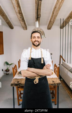 Un allegro chef maschile si trova in un ambiente accogliente e rustico, sorridendo direttamente alla telecamera. Indossa una camicia bianca pulita e una scura Foto Stock