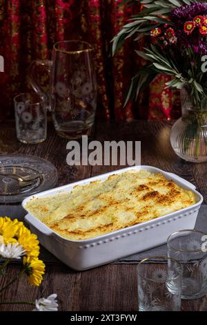 Deliziosa torta di carne fatta in casa in un piatto bianco da forno, servita su un tavolo di legno. Il piatto è circondato da eleganti bicchieri e fiori vivaci, creatina Foto Stock