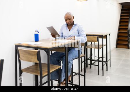Una donna concentrata siede su una scrivania di legno in una moderna classe, rivede appunti e studia con un tablet digitale accanto a lei è visibile Una bottiglia d'acqua, Foto Stock