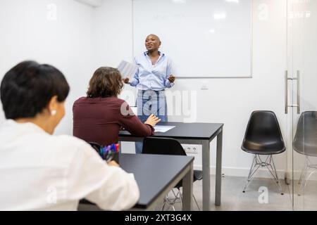 Una giovane insegnante di colore si trova davanti a un gruppo eterogeneo di studenti adulti in una classe di apprendimento inglese, dando energicamente spiegazioni e facil Foto Stock