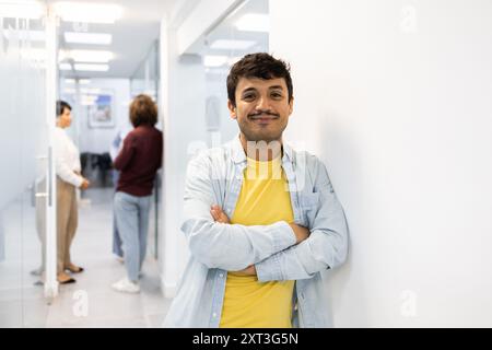 Un giovane sorride con fiducia alla telecamera, le braccia incrociate, in un corridoio ben illuminato dell'accademia inglese, con due colleghi che discutono sullo sfondo Foto Stock