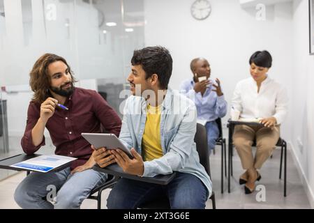 Un gruppo eterogeneo di professionisti in una classe di inglese piena di luce collabora tramite un tablet digitale due uomini discutono mentre due donne lavorano in modo indipendente Foto Stock