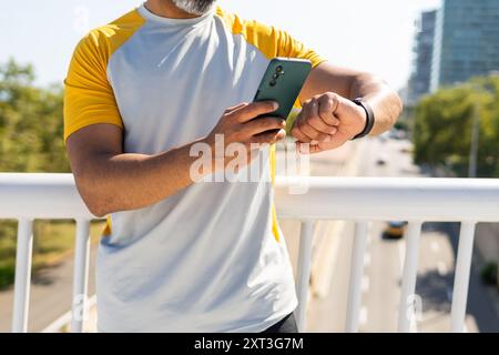 Uomo indiano dal taglio corto e irriconoscibile, vestito con un outfit estivo casual, che controlla il suo smartwatch mentre tiene in mano uno smartphone su una città Foto Stock