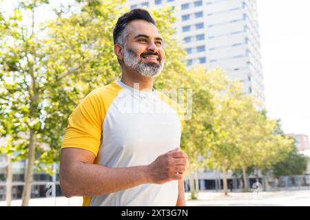 Un uomo indiano con barba chiara e capelli grigi viene visto fare jogging in un parco urbano illuminato dal sole. Indossa una camicia gialla e bianca e sorride, guardando lontano Foto Stock